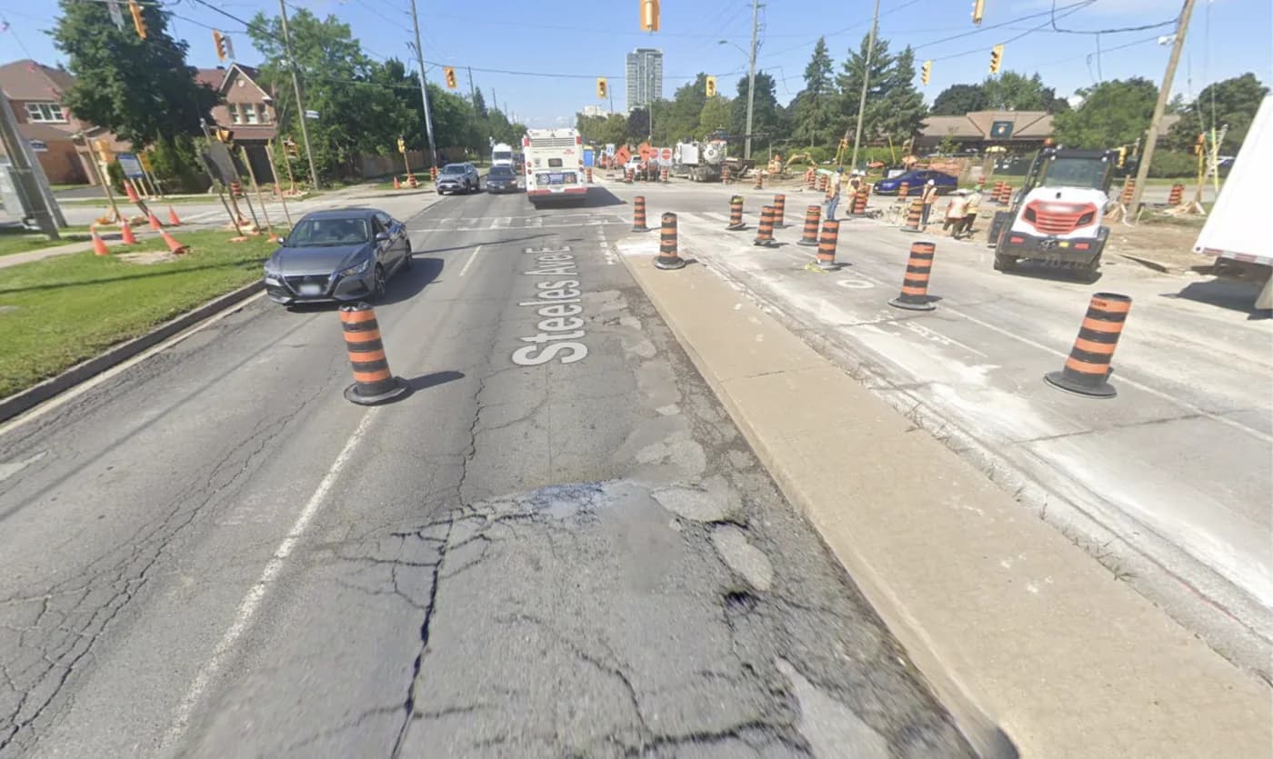 Cracked, patched pavement and construction barrels on Steeles Avenue East at Ashcott Avenue